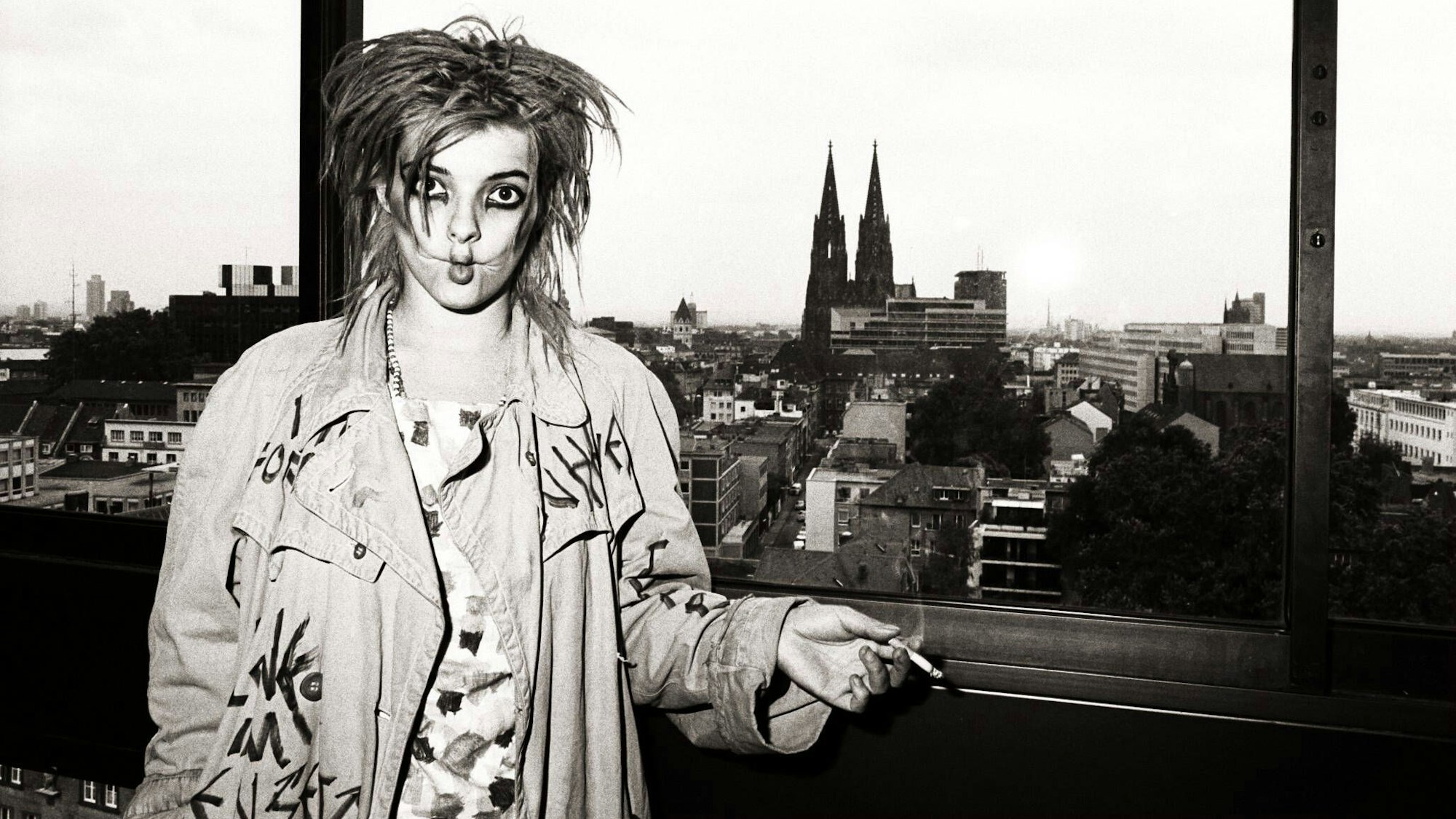 Nina Hagen raucht, den Kölner Dom im Hintergrund, eine Zigarette und zieht dabei die Wangen ein.