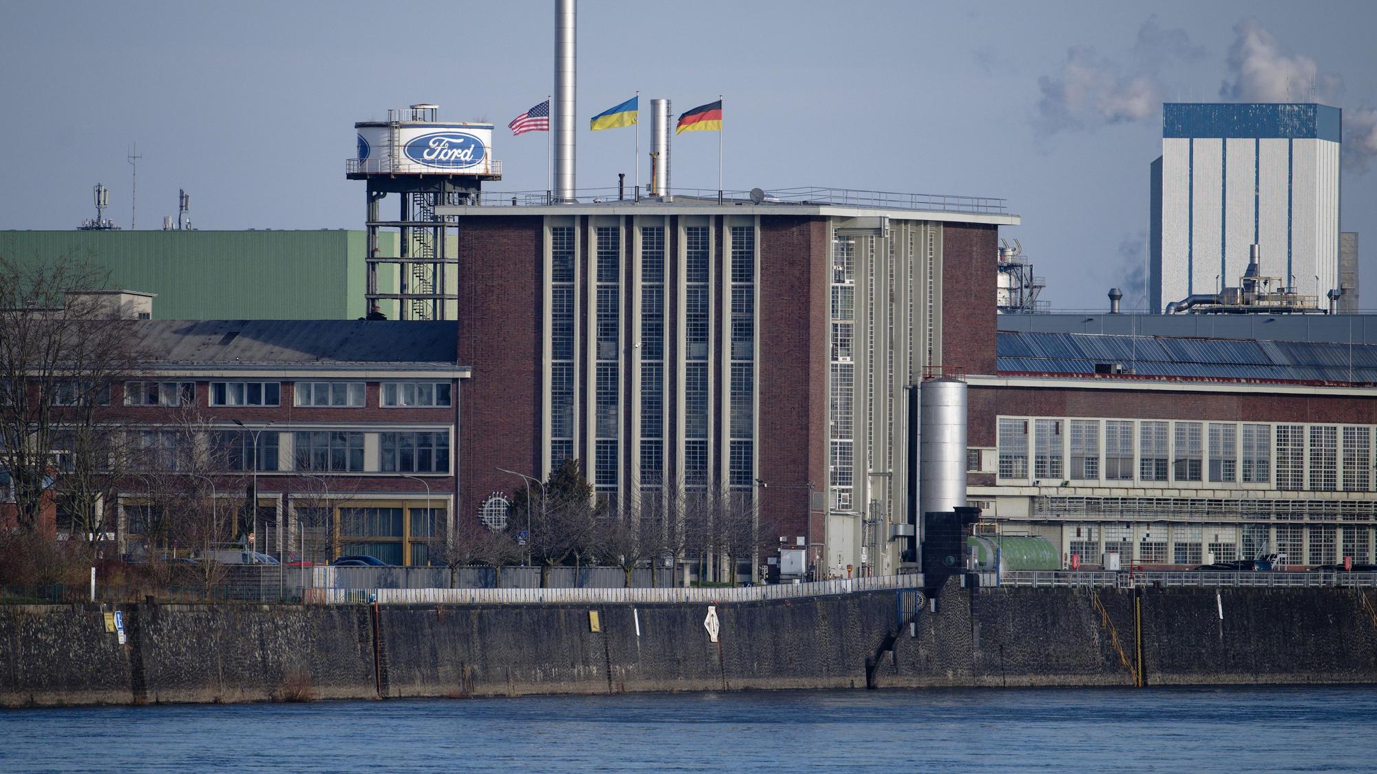 Die Flaggen der USA (l-r), der Ukraine und der Bundesrepublik Deutschland wehen über dem Dach eines Gebäudes der Fordwerke. Arbeitgeber- und Arbeitnehmervertreter sorgen sich um den Industriestandort und zigtausende daran hängende Arbeitsplätze in Nordrhein-Westfalen.