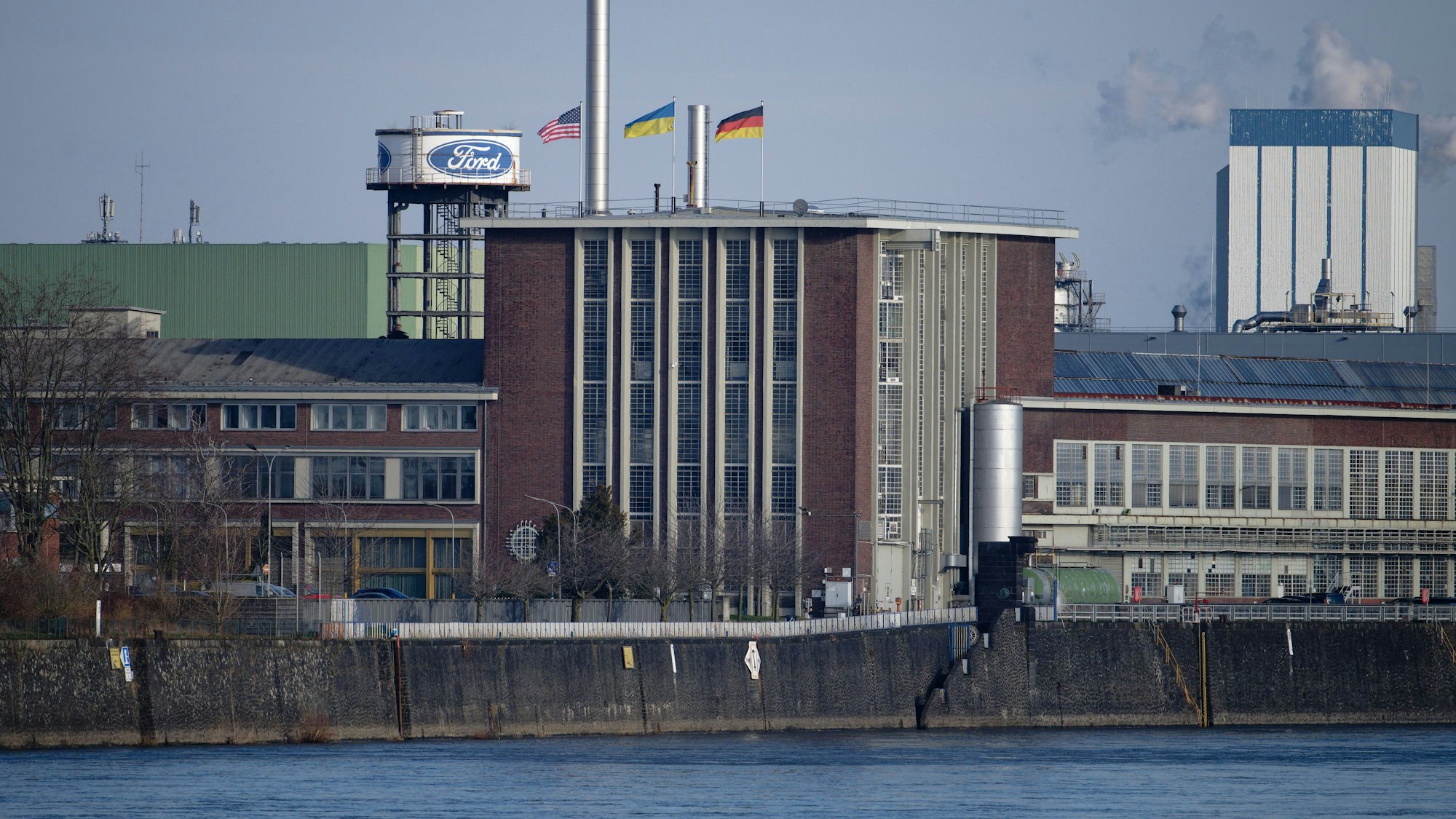 Die Flaggen der USA (l-r), der Ukraine und der Bundesrepublik Deutschland wehen über dem Dach eines Gebäudes der Fordwerke. Arbeitgeber- und Arbeitnehmervertreter sorgen sich um den Industriestandort und zigtausende daran hängende Arbeitsplätze in Nordrhein-Westfalen.