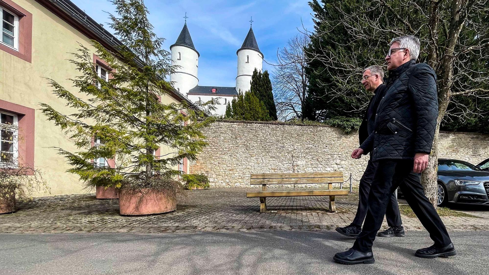 Zwei in schwarz gekleidete Männer gehen in Kall-Steinfeld einen Weg entlang. Im Hintergrund sind die Türme der Basilika zu sehen.