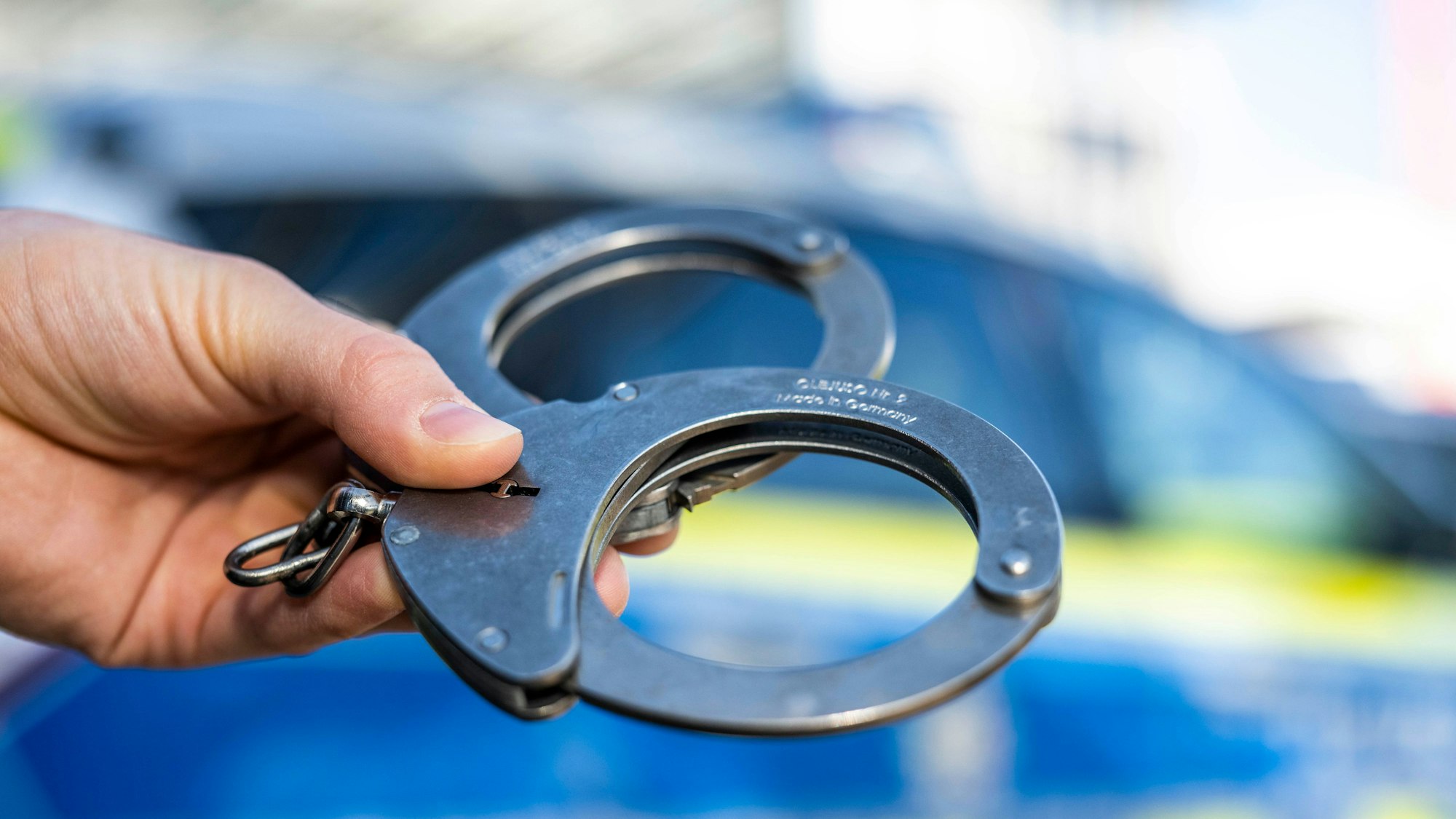 Eine Hand hält Handschellen vor einen Streifenwagen der Polizei.
