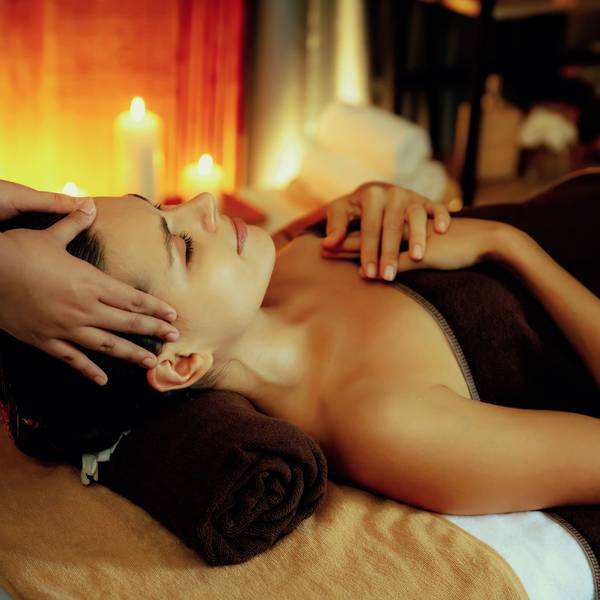 Eine Frau liegt auf einer Massageliege, es sind Kerzen an und die Frau wird am Kopf massiert.