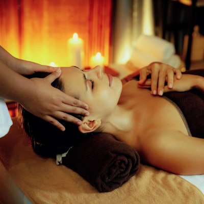 Eine Frau liegt auf einer Massageliege, es sind Kerzen an und die Frau wird am Kopf massiert.