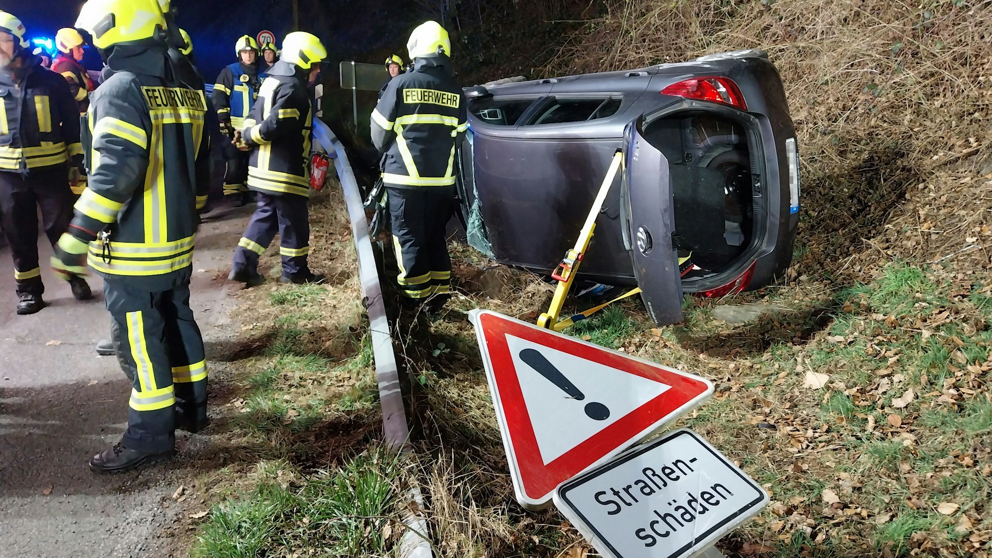 Ein Auto liegt am Straßenrand auf der Seite. Links Feuerwehrleute. Im Vordergrund ein Straßenschild mit der Aufschrift „Straßenschäden“.