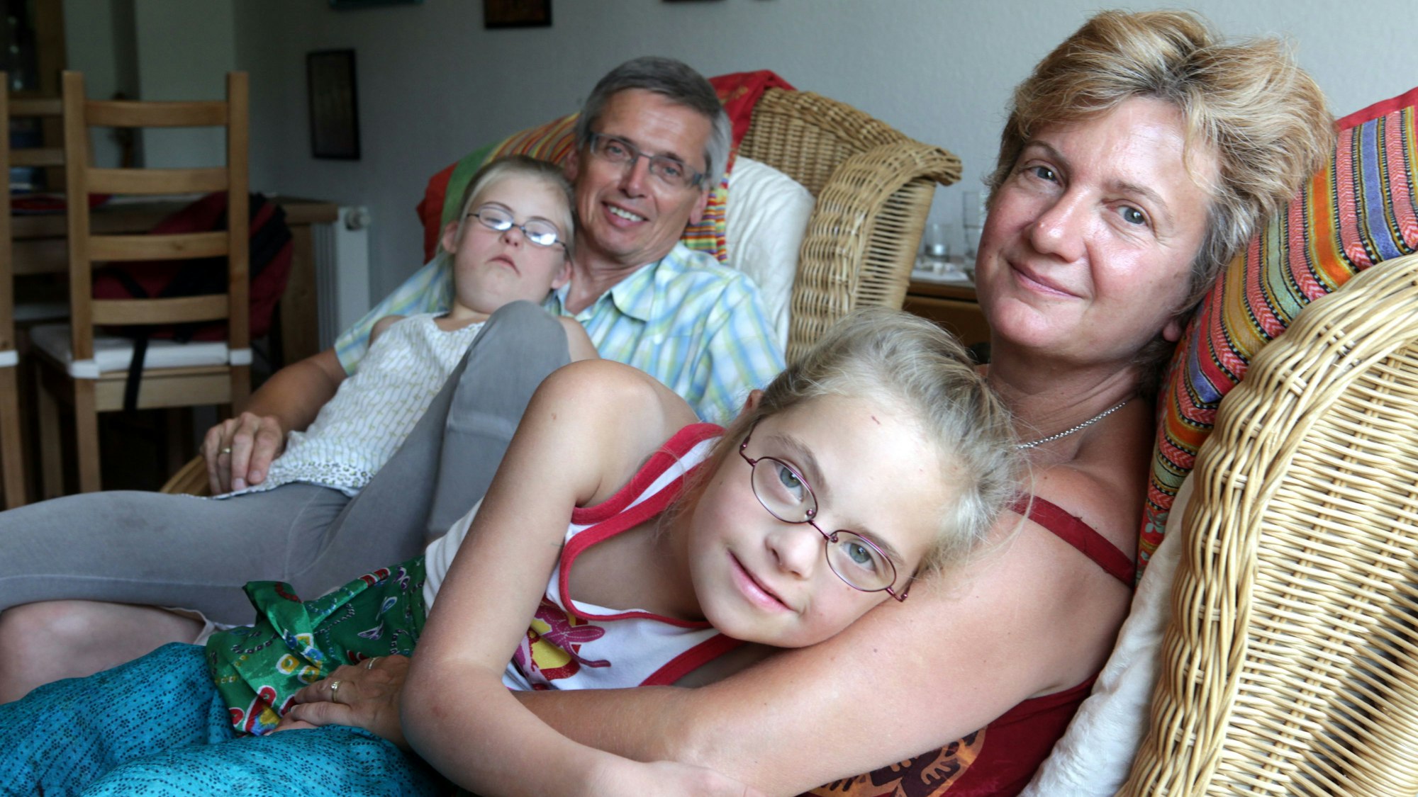 Ein Mann und eine Frau sitzen auf Rattan-Korbstühlen. Beide haben jeweils ein Mädchen mit blonden Haaren und Brille auf dem Schoß.