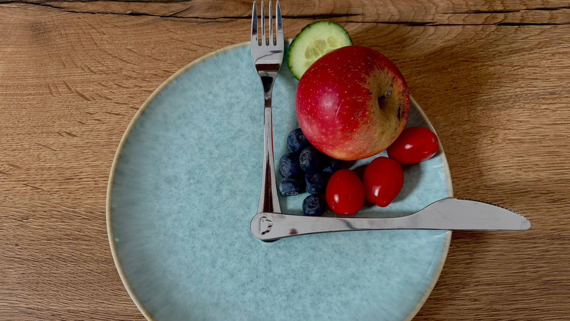 Ein Teller, darauf Messer und Gabel wie Uhrzeiger, dazwischen gesundes Essen, etwa Tomaten, Gurke, Beeren und Apfel.