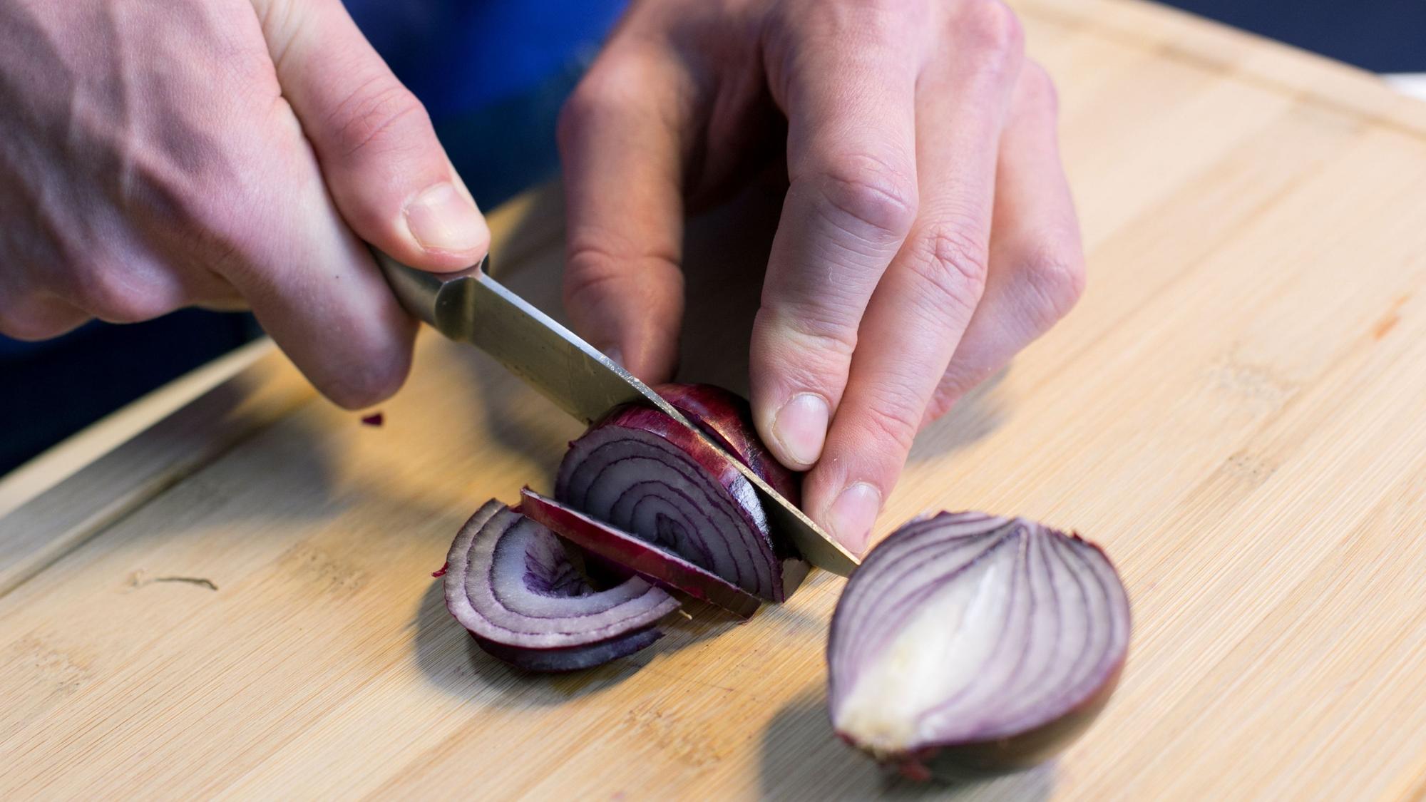 Zu sehen: Zwei Hände, die eine hält ein Messer, die andere eine rote Zwiebel, die mit dem scharfen Messer geschnitten wird.
