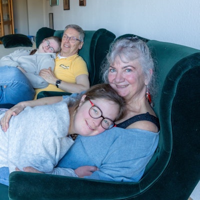 In einem Wohnzimmer sitzen auf zwei grünen Sesseln ein Mann und eine Frau. Beide haben jeweils eine junge Frau auf dem Schoß.