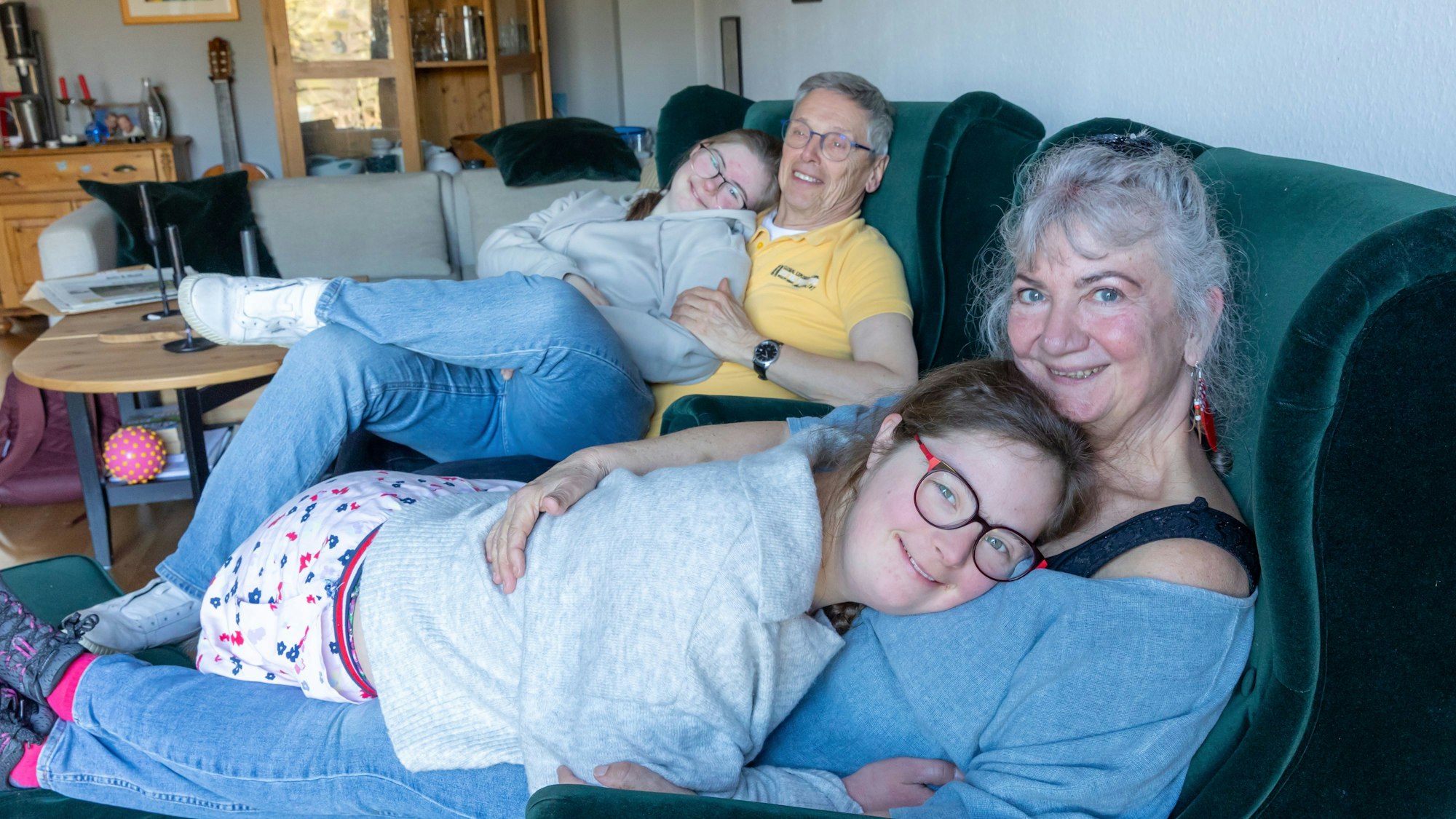 In einem Wohnzimmer sitzen auf zwei grünen Sesseln ein Mann und eine Frau. Beide haben jeweils eine junge Frau auf dem Schoß.