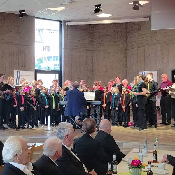 Kinder, Frauen und Männer singen gemeinsam auf einer Bühne.