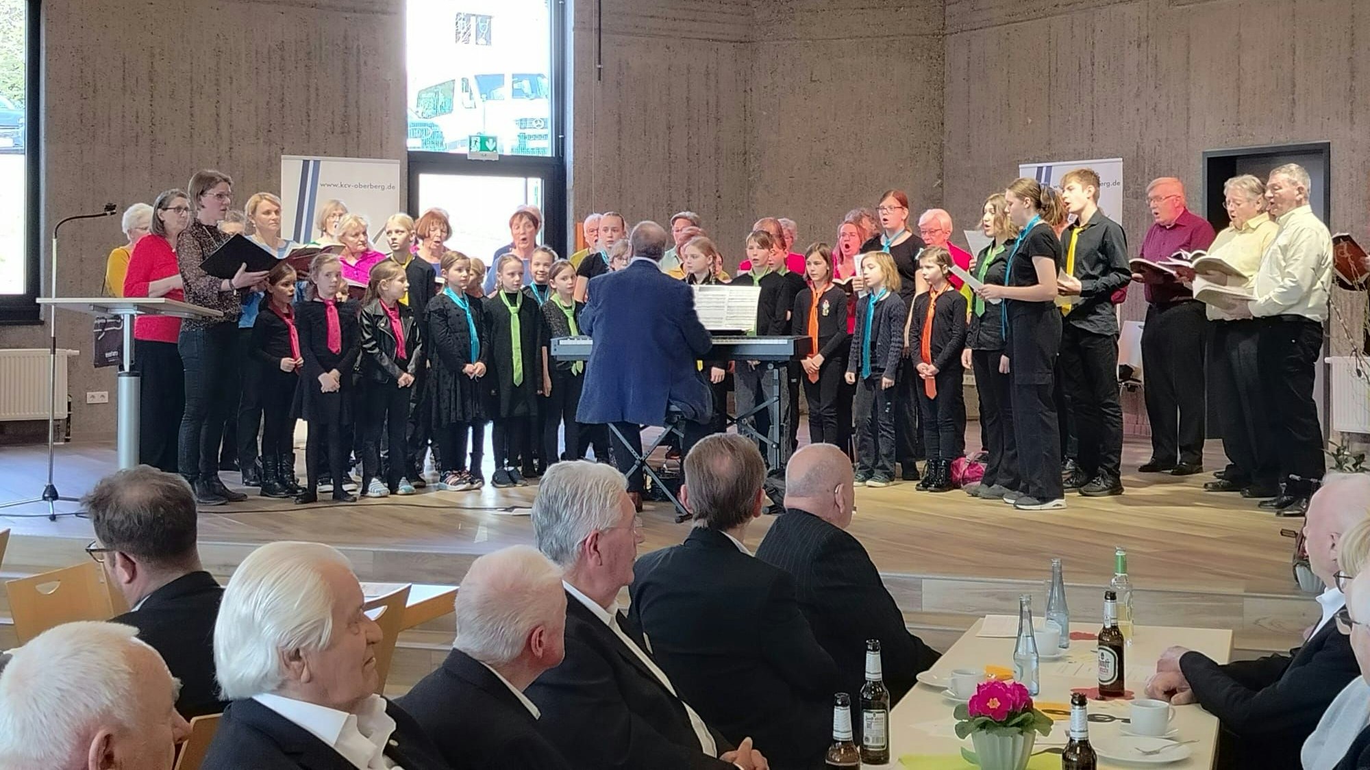 Kinder, Frauen und Männer singen gemeinsam auf einer Bühne.