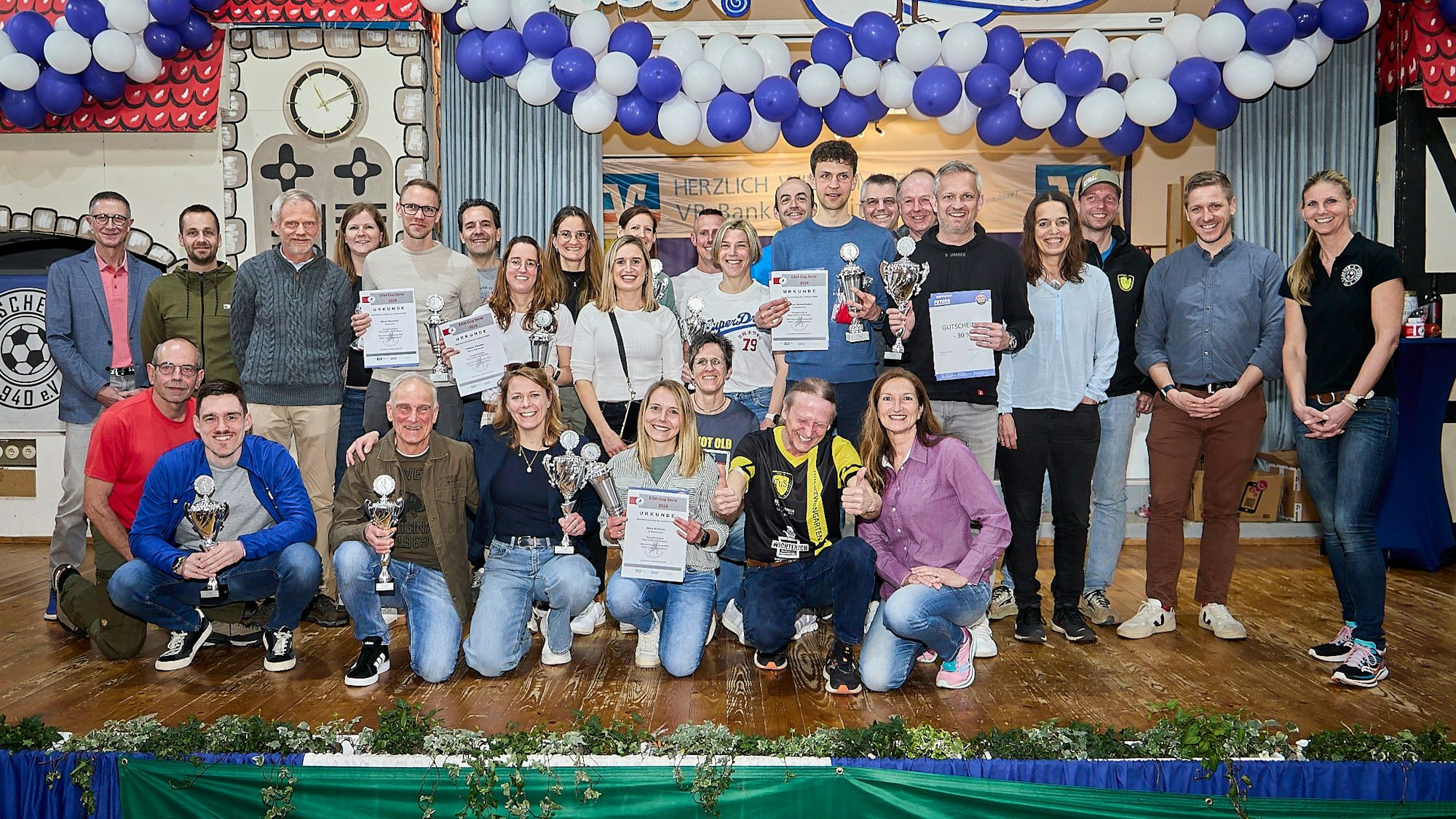 Zahlreiche Frauen und Männer stehen und hocken auf der Bühne im Dorfsaal in Scheven. Sie halten Pokale und Urkunden in den Händen, die sie beim Eifelcup gewonnen haben.