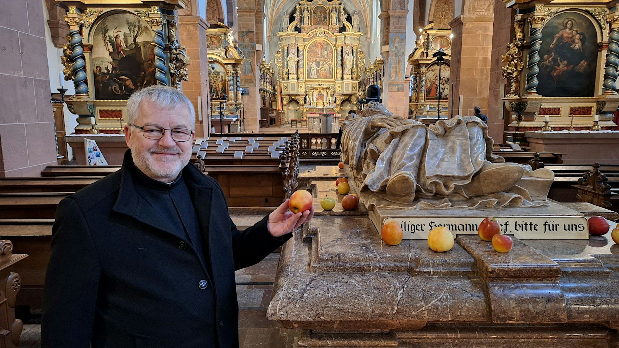 Ein Mann in dunkler Kleidung steht neben einem Sarkophag in einer Kirche und hält einen Apfel in der Hand.