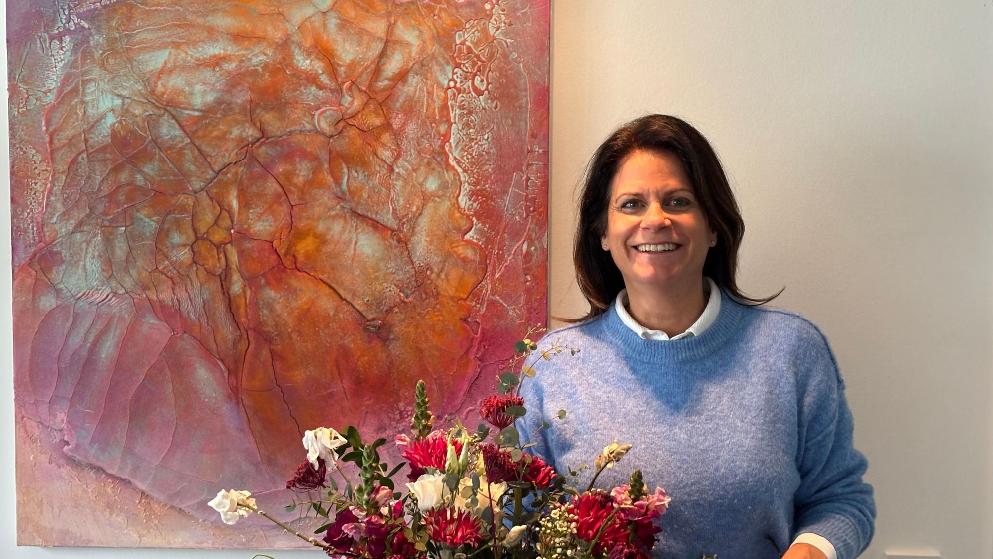 Unten mittig im Bild steht ein Blumenstrauß. Dahinter steht eine Frau neben einem Bild, das an der Wand hängt, und lächelt in die Kamera.