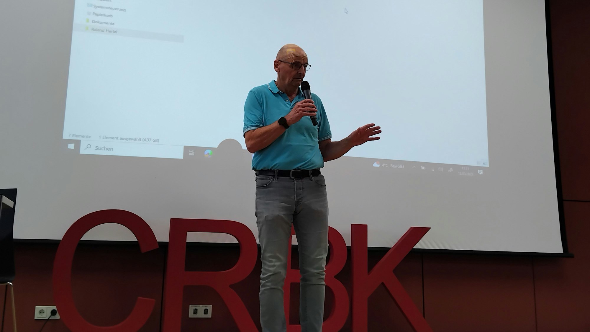 Ein Mann steht auf einer Bühne und spricht gestikulierend in ein Mikrophon.