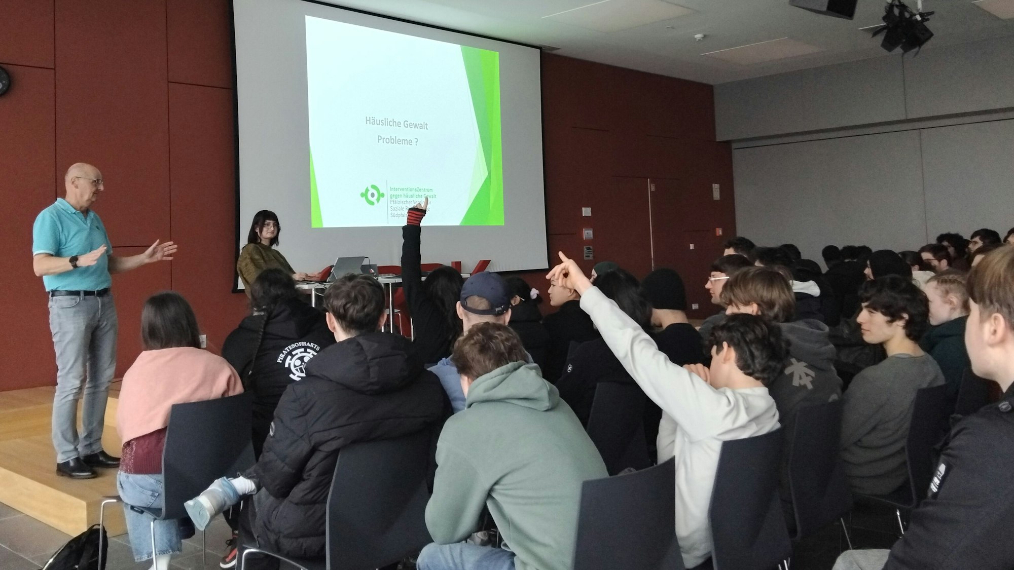 Eine Gruppe Jugendlicher und junger Erwachsener sitzt in einer Aula, zwei Leute haben die Hände gehoben. Ein älterer Mann steht gestikulierend vor ihnen. Auf einer Leinwand vorne steht „Häusliche Gewalt - Probleme?“