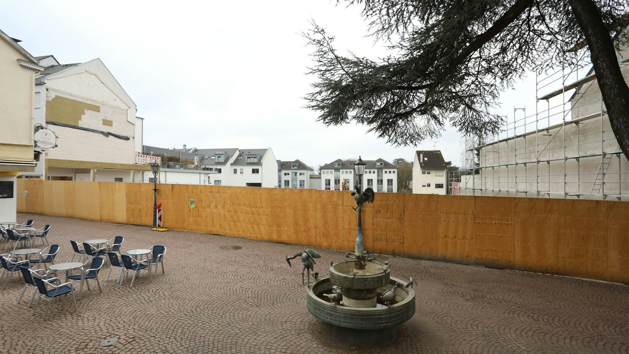 Ein Brunnen und einige Tische und Stühle stehen in der Fußgängerzone, im Hintergrund steht ein großer Bauzaun.