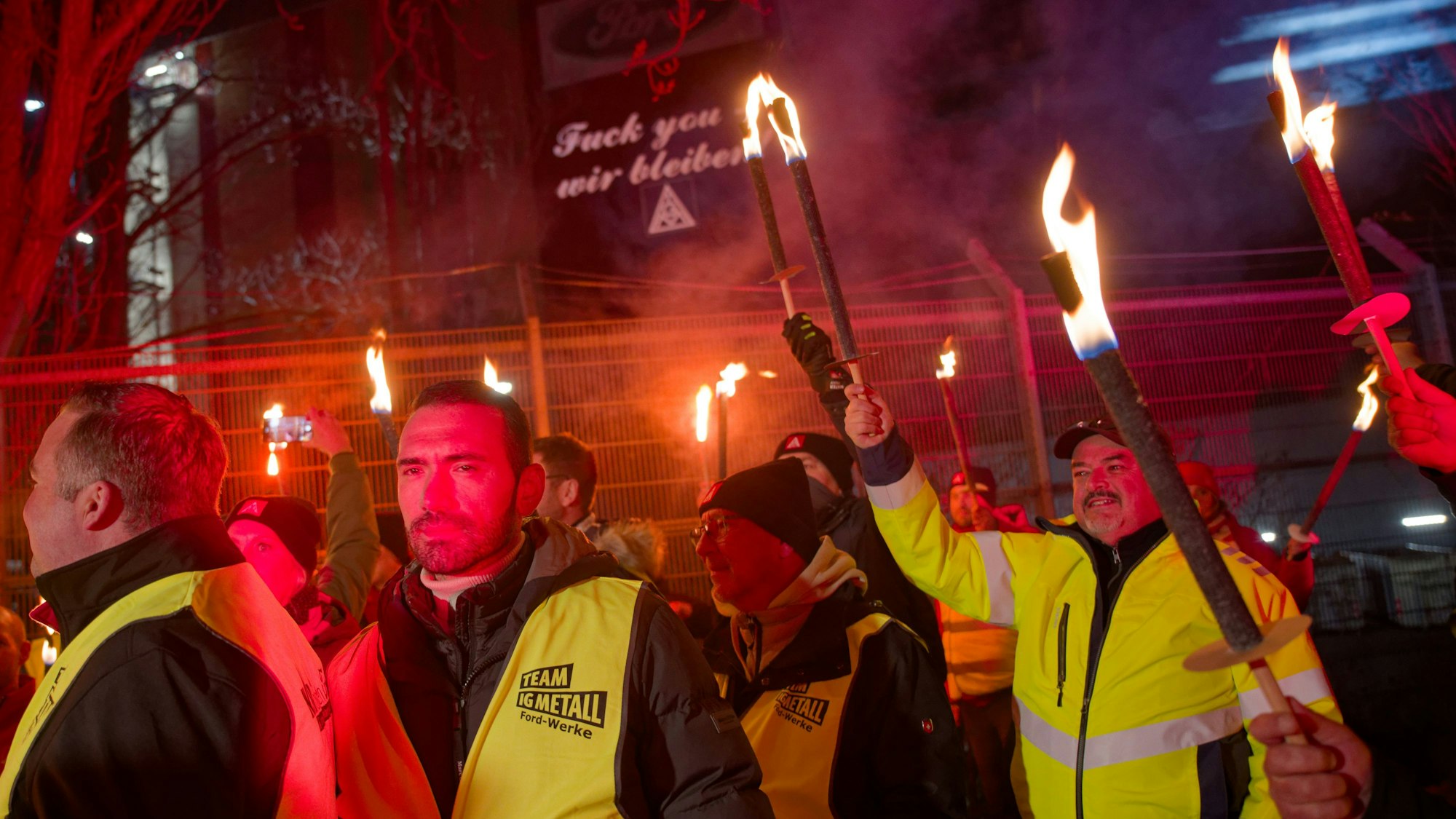 11.03.2025, Nordrhein-Westfalen, Köln: Ford-Mitarbeiter stehen mit Fackeln auf dem Gehweg vor einem Gebäude der Fordwerke, auf dem die Worte "Fuck you - wir bleiben" projiziert wird. Foto: Henning Kaiser/dpa +++ dpa-Bildfunk +++
