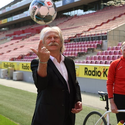Henning Krautmacher und Oliver Gritz stehen im RheinEnergiestadion, Krautmacher balanciert einen Ball mit dem Finger und Oliver Gritz steht in einem Fahrradtrikot und einem Rennrad dahinter.