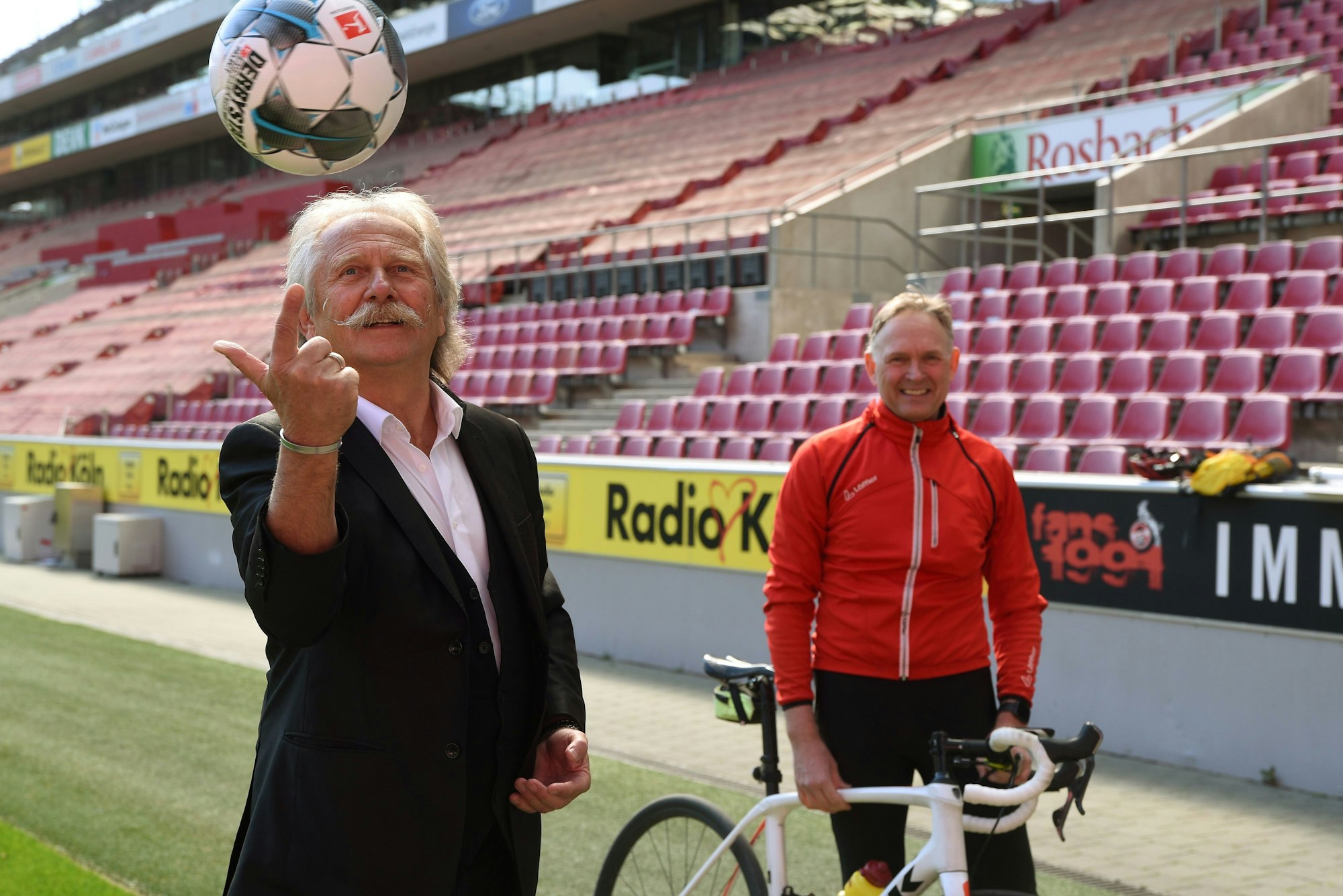 Die Initiatoren des Kölner Benefiz-Leselaufs im und ums Rhein-Energie-Stadion: Henning Krautmacher und Oliver Gritz stehen im Stadion. Henning Krautmacher jongliert einen Fußball, Oliver Gritz hält sein Rennrad in der Hand.