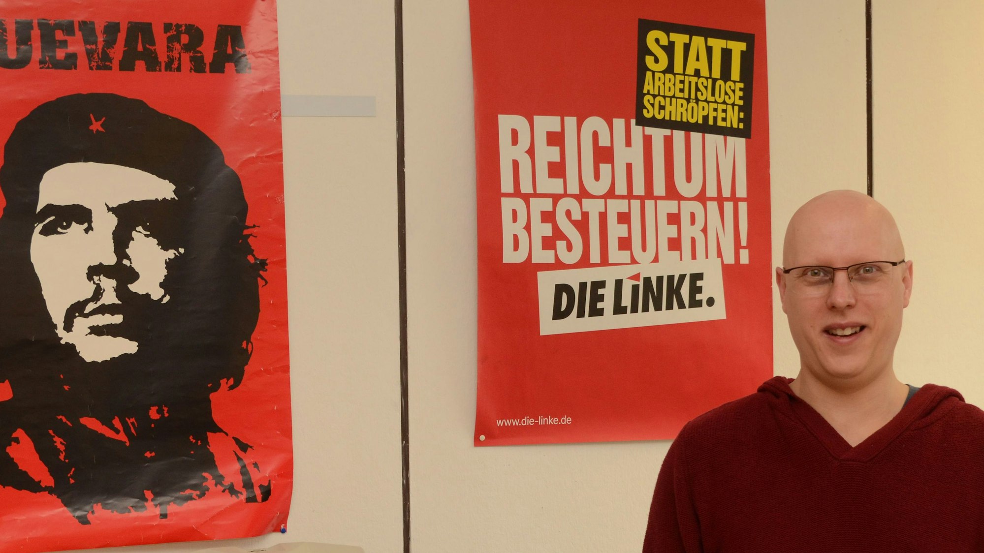 Ein Mann mit Glatze steht vor einer Wand mit politischen Plakaten der Partei Die Linke.