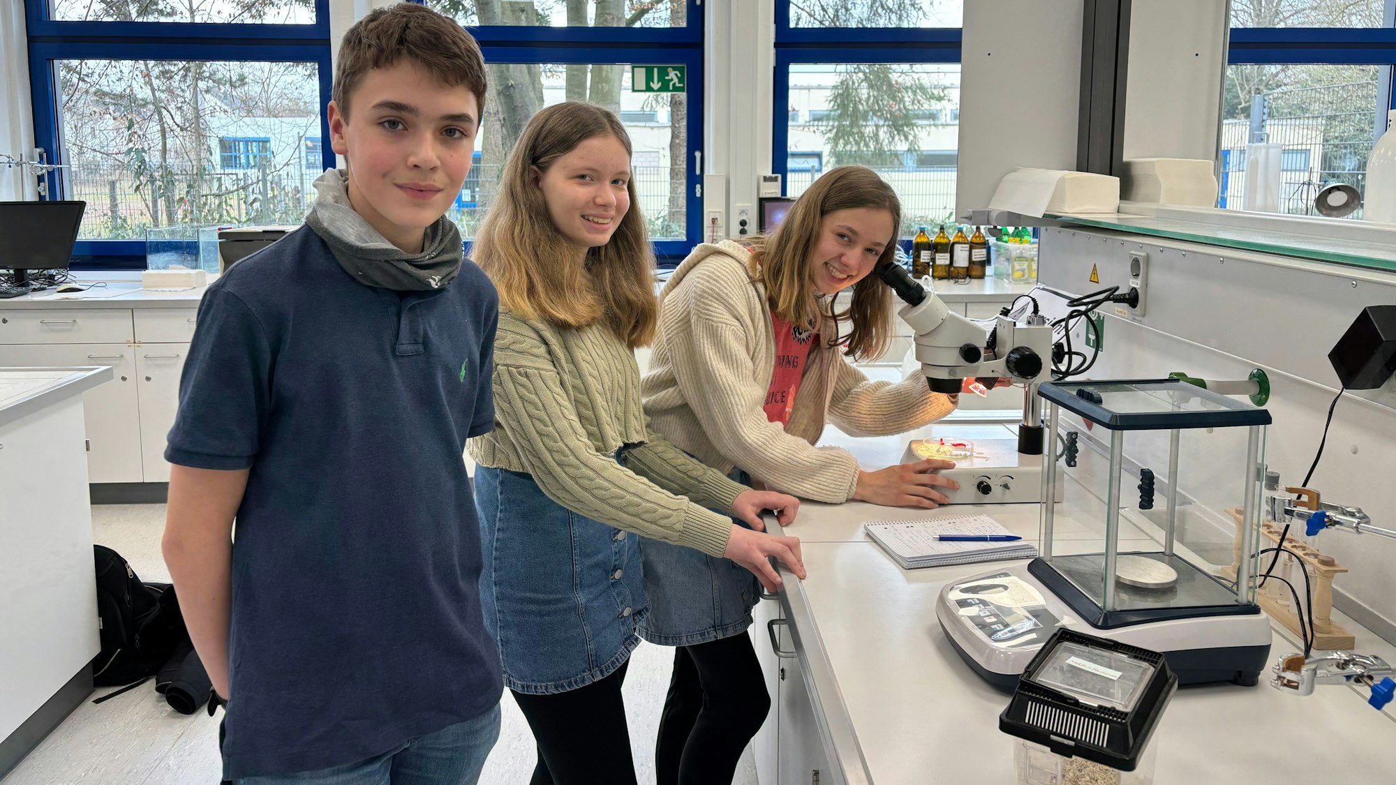 Ein Junge und zwei Mädchen stehen in einem Labor, in dem ein Behälter, eine Waage und ein Mikroskop stehen.