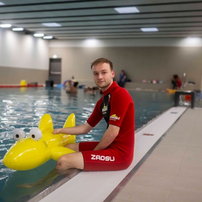 Ein Schwimmlehrer in rotem Neoprenanzug sitzt am Rand des Schwimmbackens der Schwimmschule Sharky in Weilerswist. Die Beine baumeln im Wasser. Er hält einen gelben Plastik-Hai fest.