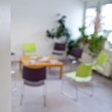 Das Foto gibt den Blick frei in ein leeres Wartezimmer in einer Arztpraxis. Auf der geöffneten Tür befindet sich der Aufdruck „Wartezimmer“, im Raum sind Stühle, ein niedriger Tisch mit Lesematerial sowie Grünpflanzen zu sehen.