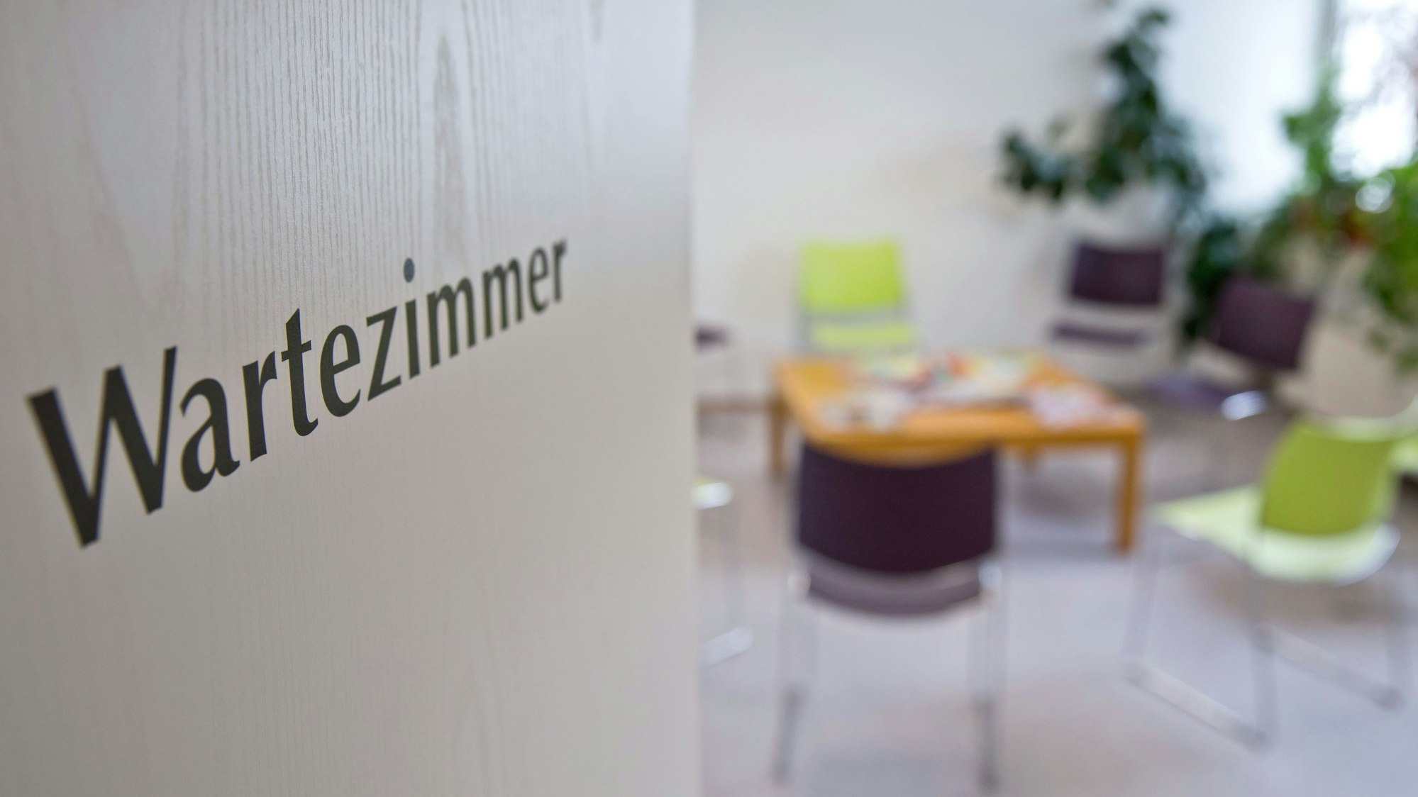 Das Foto gibt den Blick frei in ein leeres Wartezimmer in einer Arztpraxis. Auf der geöffneten Tür befindet sich der Aufdruck „Wartezimmer“, im Raum sind Stühle, ein niedriger Tisch mit Lesematerial sowie Grünpflanzen zu sehen.