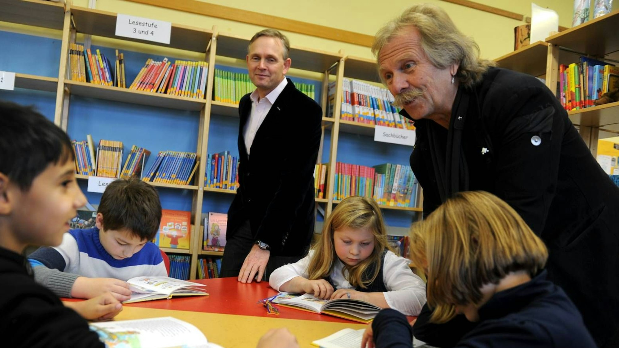 Henning Krautmacher und Oliver Gritz stehen bei der Eröffnung einer der ersten Leseclubs im Jahr 2012 in der KGS Heßhofstraße in Vingst an einem Tisch, an dem vier Grundschülerinnen und Grundschüler in einem Buch lesen.
