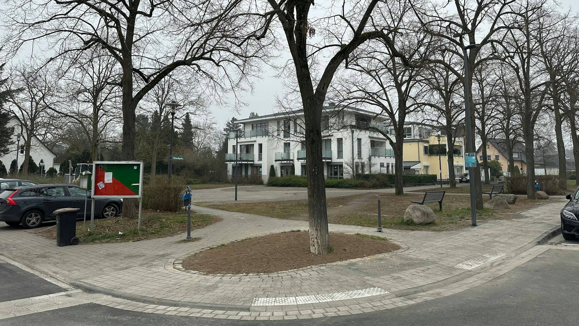 Ein städtischer Platz, ringsum stehen Bäume, im Hintergrund sind Wohnhäuser zu sehen.