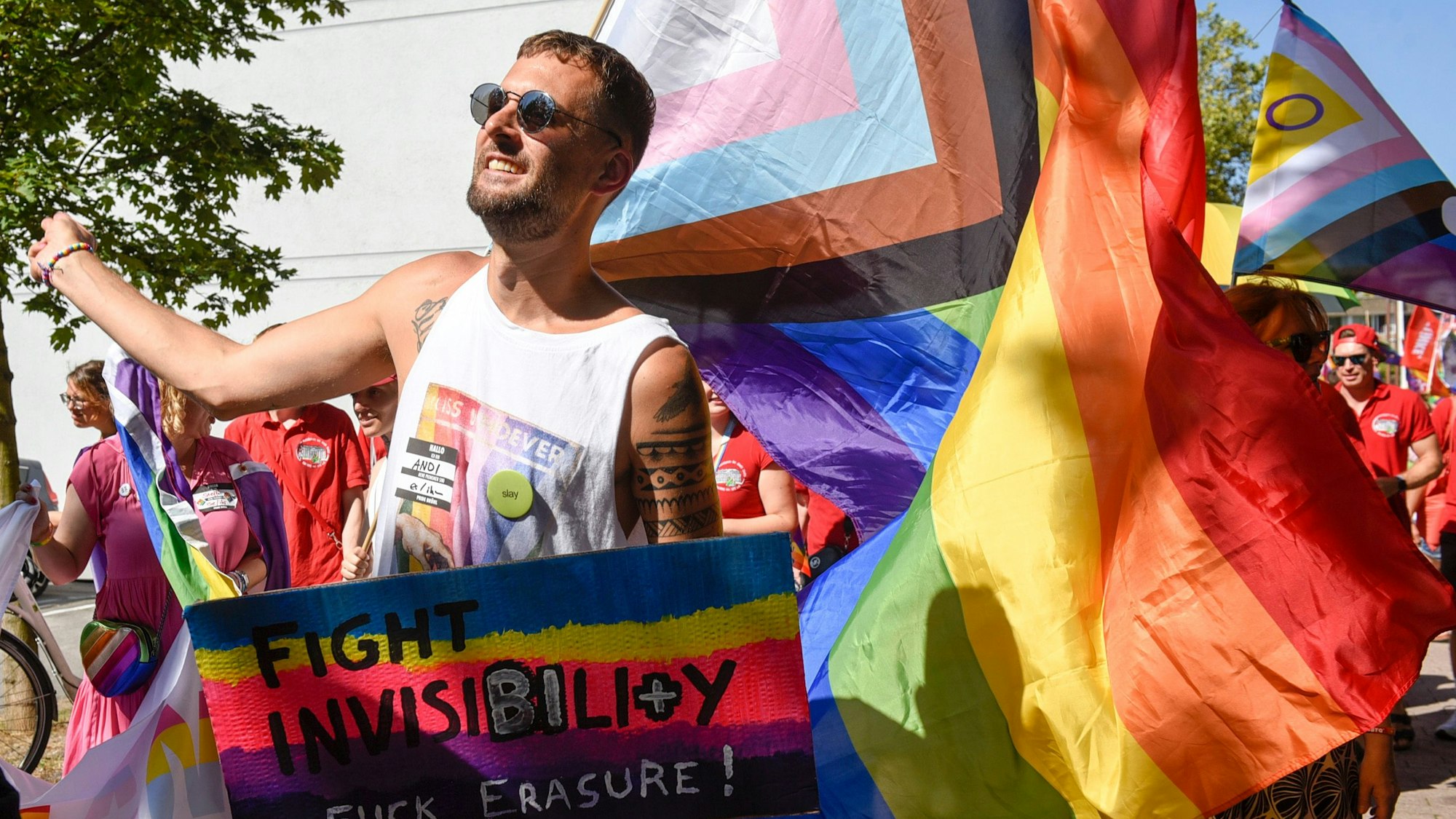 Eine Person hält ein Schild mit der Aufschrift Fight Invisibility in der Hand. Hinter der Person wehen Pride-Flaggen.