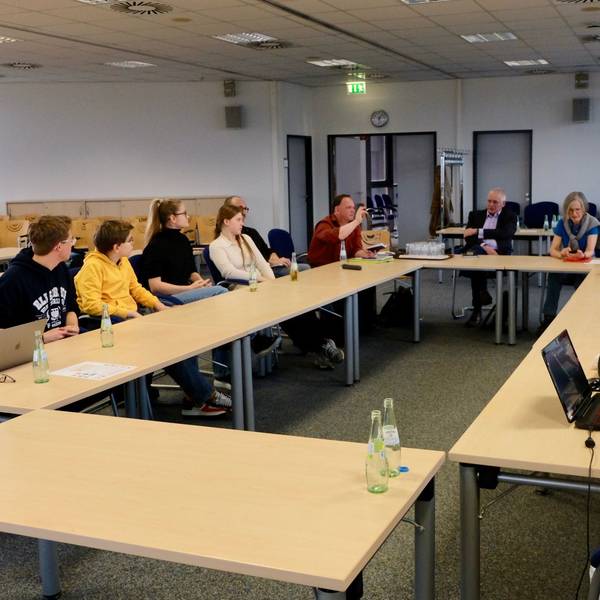 Jugendliche und Erwachsene sitzen in einem Sitzungssaal an Tischen zusammen.