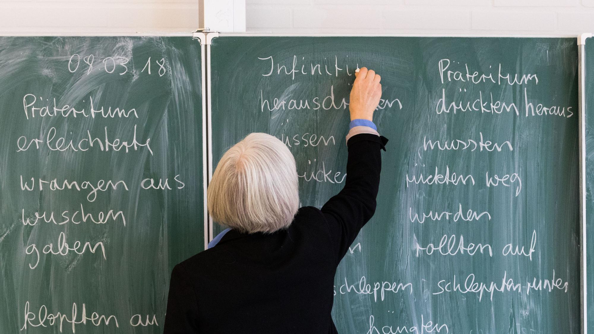 Eine Lehrerin steht in einem Klassenraum an einer Tafel und schreibt.