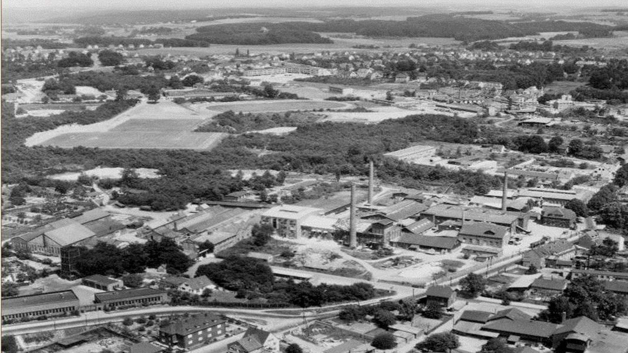 Auf einem Schwarz-weiß-Luftbild ist eine Fabrik zu sehen, an die auf der einen Seite Grünflächen, auf der anderen Wohn- und Gewerbebauung anschließen.