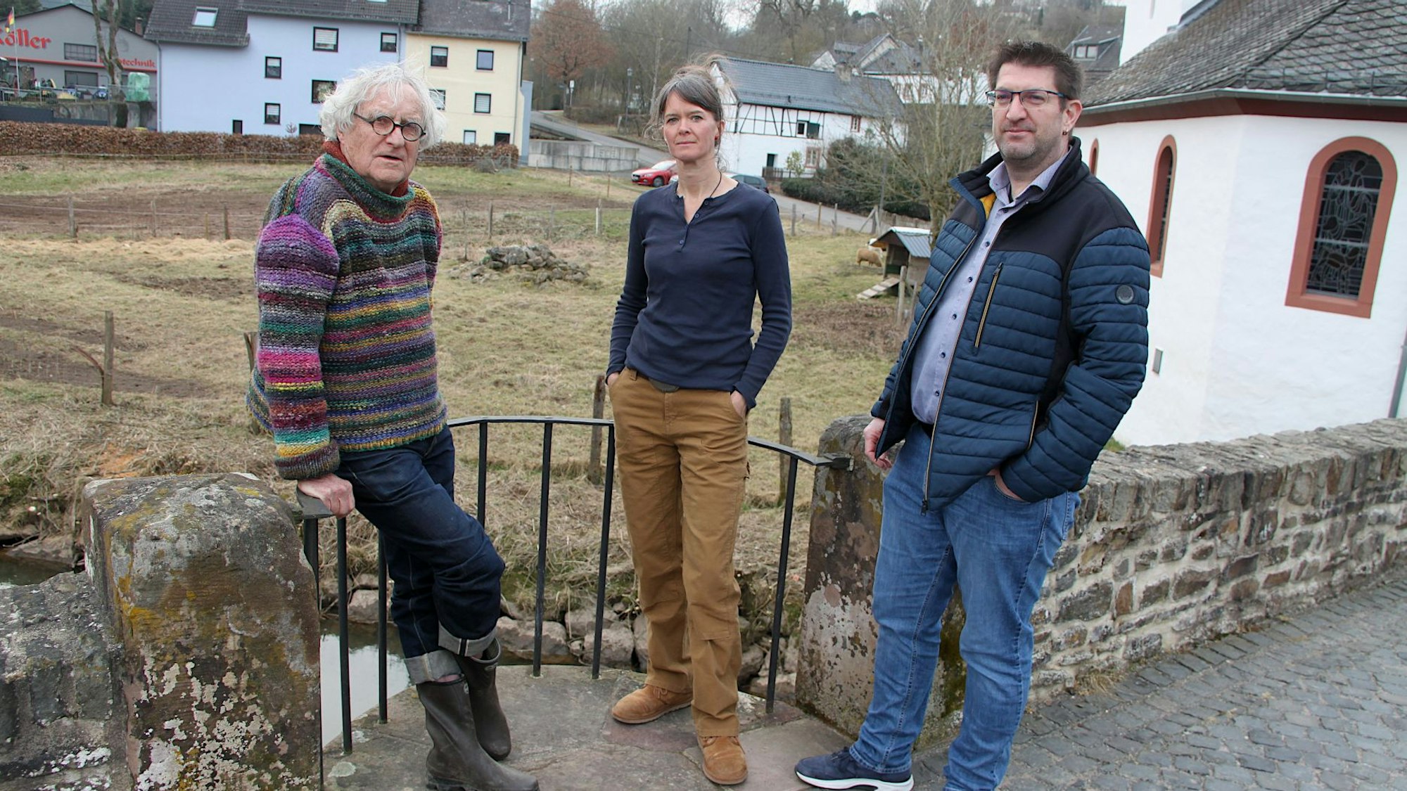 Zwei Männer und eine Frau stehen an einer alten Bruchsteinmauer, im Hintergrund sind Wiesen und Häuser zu sehen.