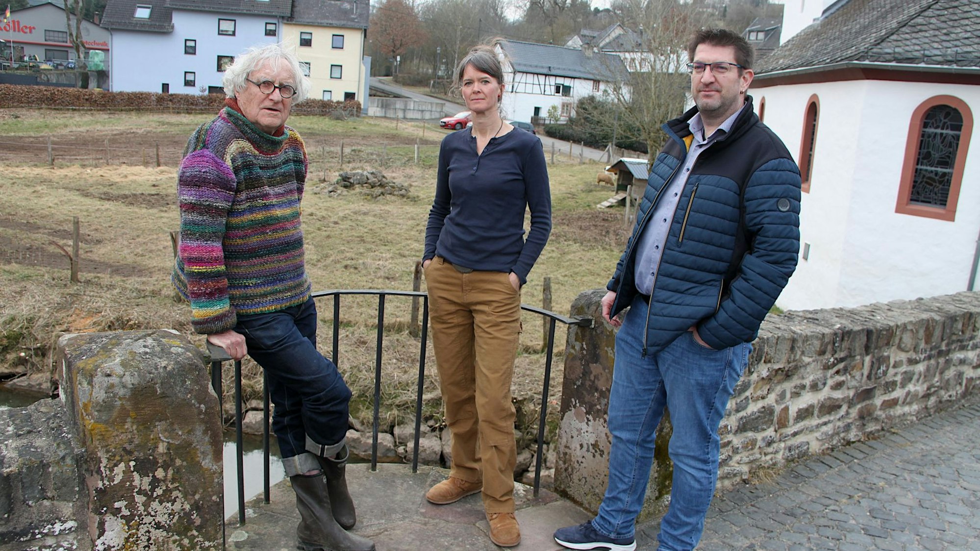 Zwei Männer und eine Frau stehen an einer alten Bruchsteinmauer, im Hintergrund sind Wiesen und Häuser zu sehen.