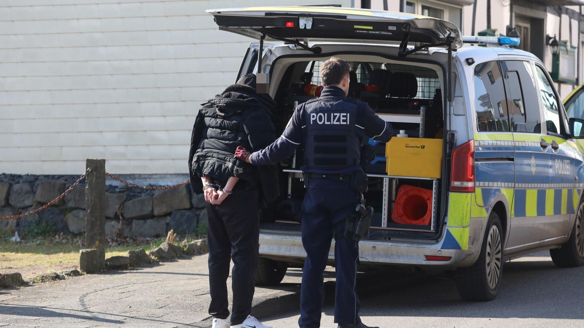 Ein Mann, dessen Hände mit Handschellen auf dem Rücken gefesselt sind, steht neben einem Polizisten hinter einem Streifenwagen.