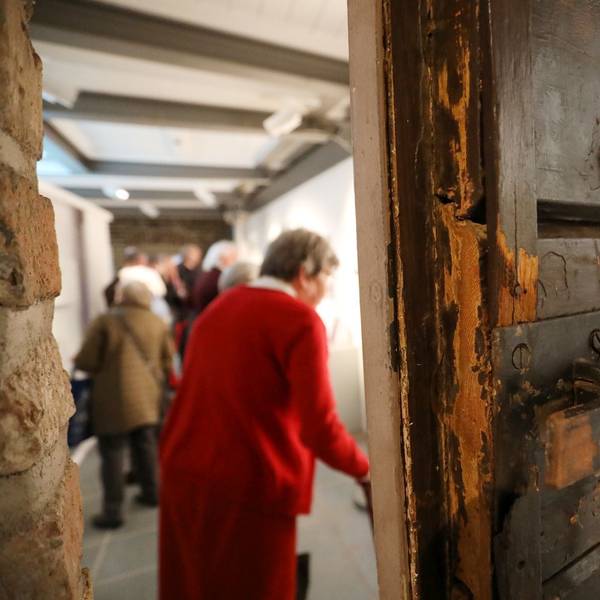 Hinter einer schweren alten Holztür, die halb geöffnet ist, stehen Menschen in einem Raum.
