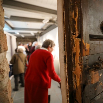 Hinter einer schweren alten Holztür, die halb geöffnet ist, stehen Menschen in einem Raum.