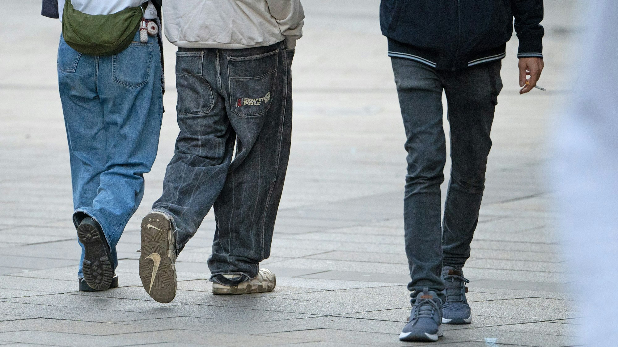 Drei Jugendliche in Jeans gehen durch die Innenstadt. Einer hält eine Zigarette in der Hand.