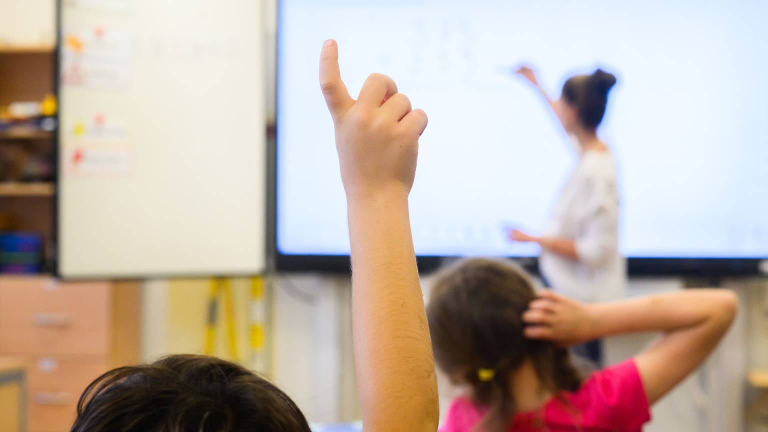 Ein Schüler meldet sich per Handzeichen, während eine Lehrerin auf eine digitale Schultafel einer Grundschule schreibt.