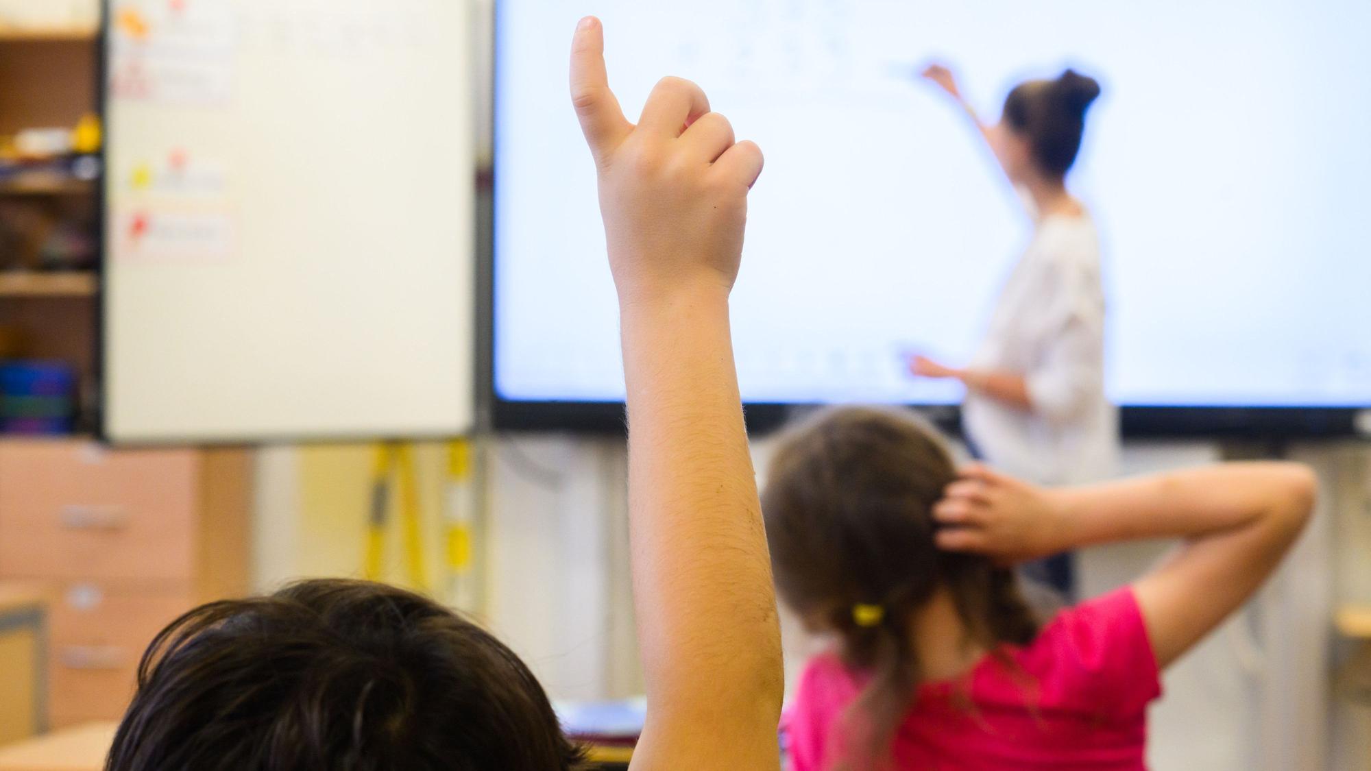 Ein Schüler meldet sich per Handzeichen, während eine Lehrerin auf eine digitale Schultafel einer Grundschule schreibt.