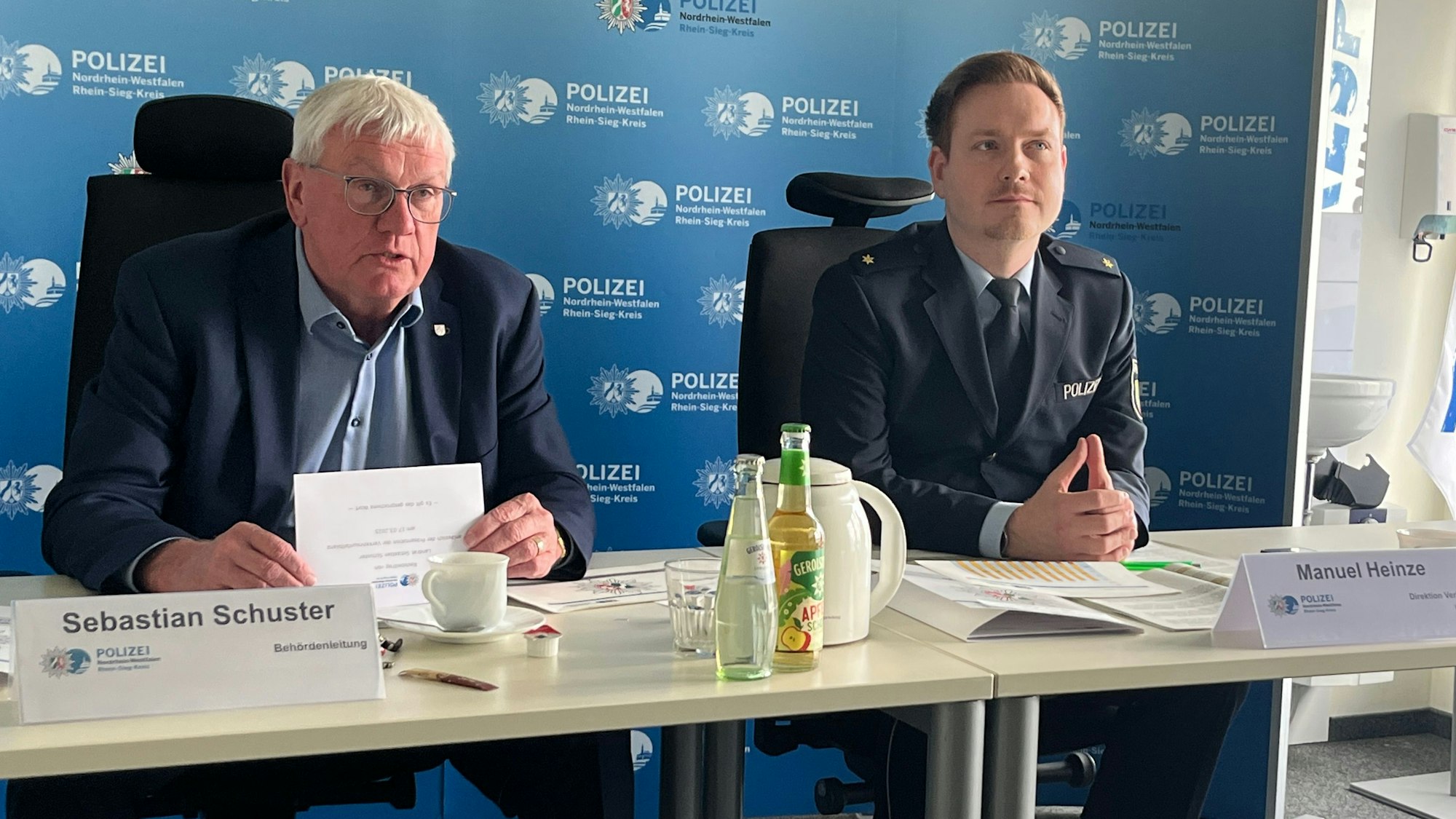 Zwei Männer sitzen vor einer blauen Wand mit Polizeiaufschriften.