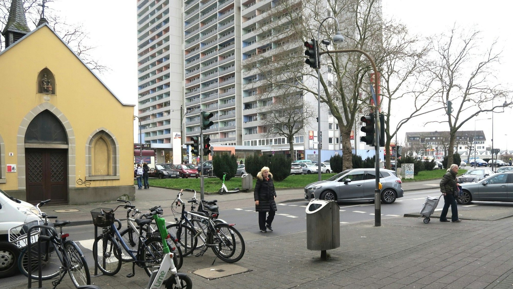 Fußgänger gehen über die Straße, Autos warten bis sie vorbei sind. Im Hintergrund sind Rochuskapelle und ein Hochhaus zu sehen.