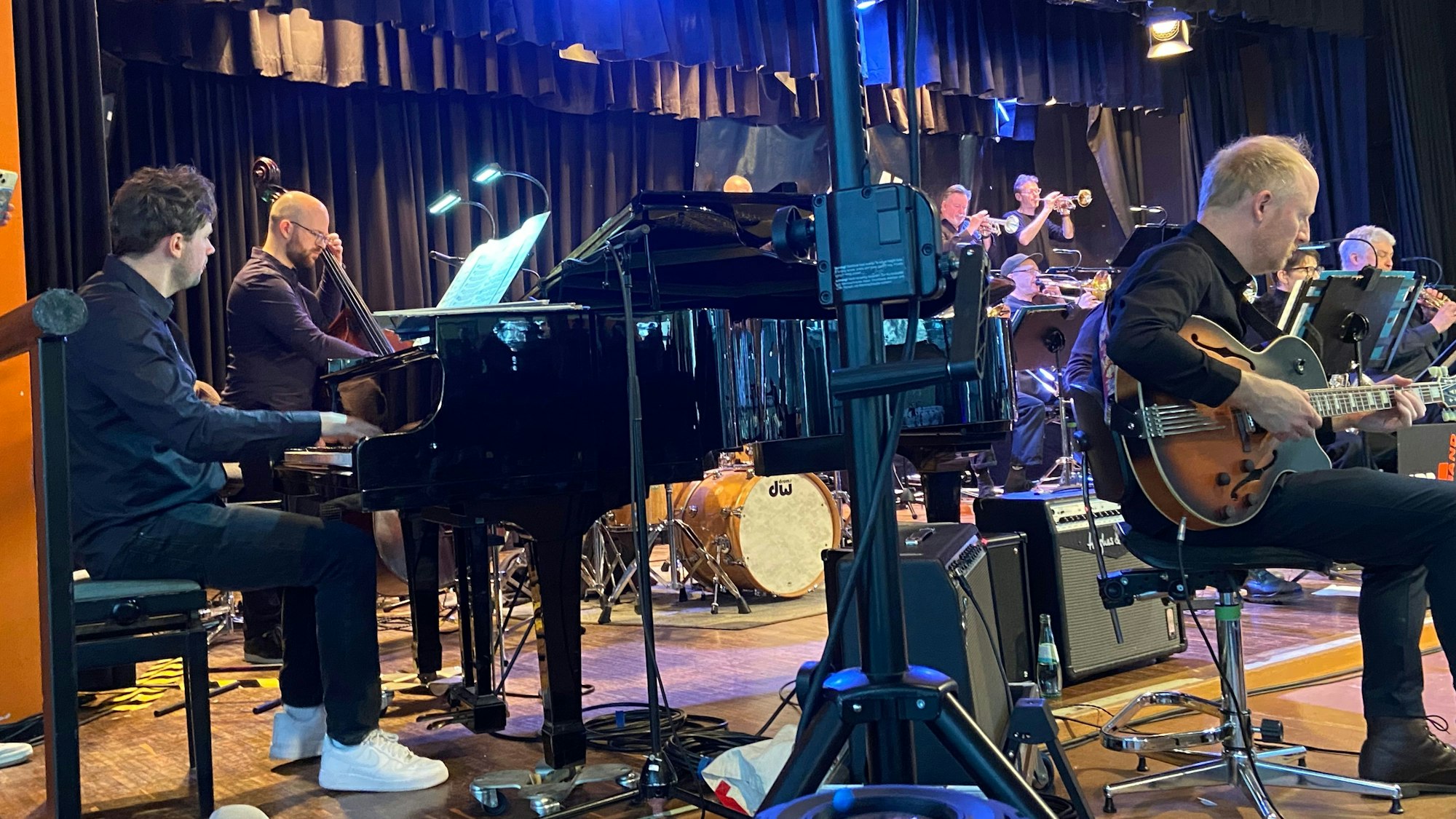 Musiker mit Kontrabass, Trompeten und Gitarre sind zu sehen. Im Vordergrund sitzt ein junger Mann am Klavier.