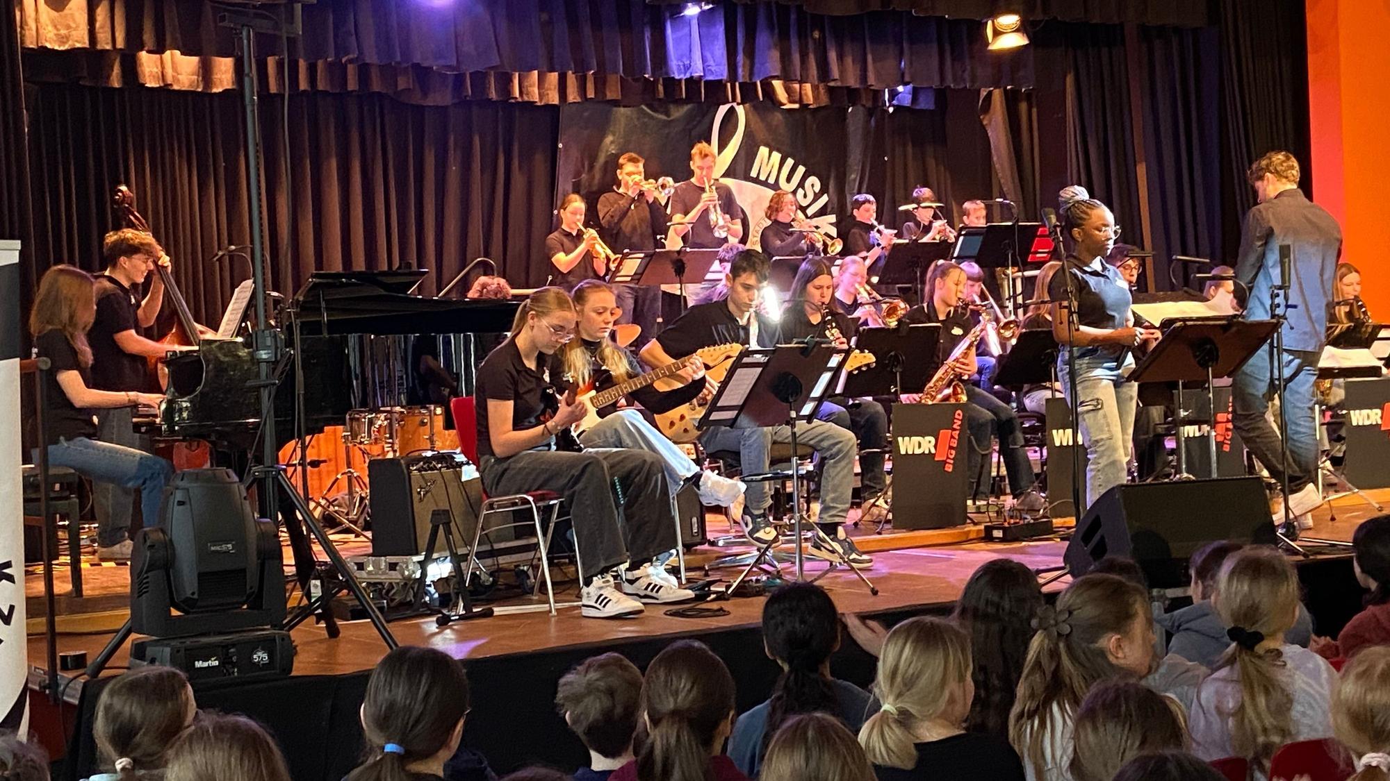 Auf einer Bühne ist ein Orchester zu sehen, im Publikum sitzen viele Schüler des Porzer Stadtgymnasiums.