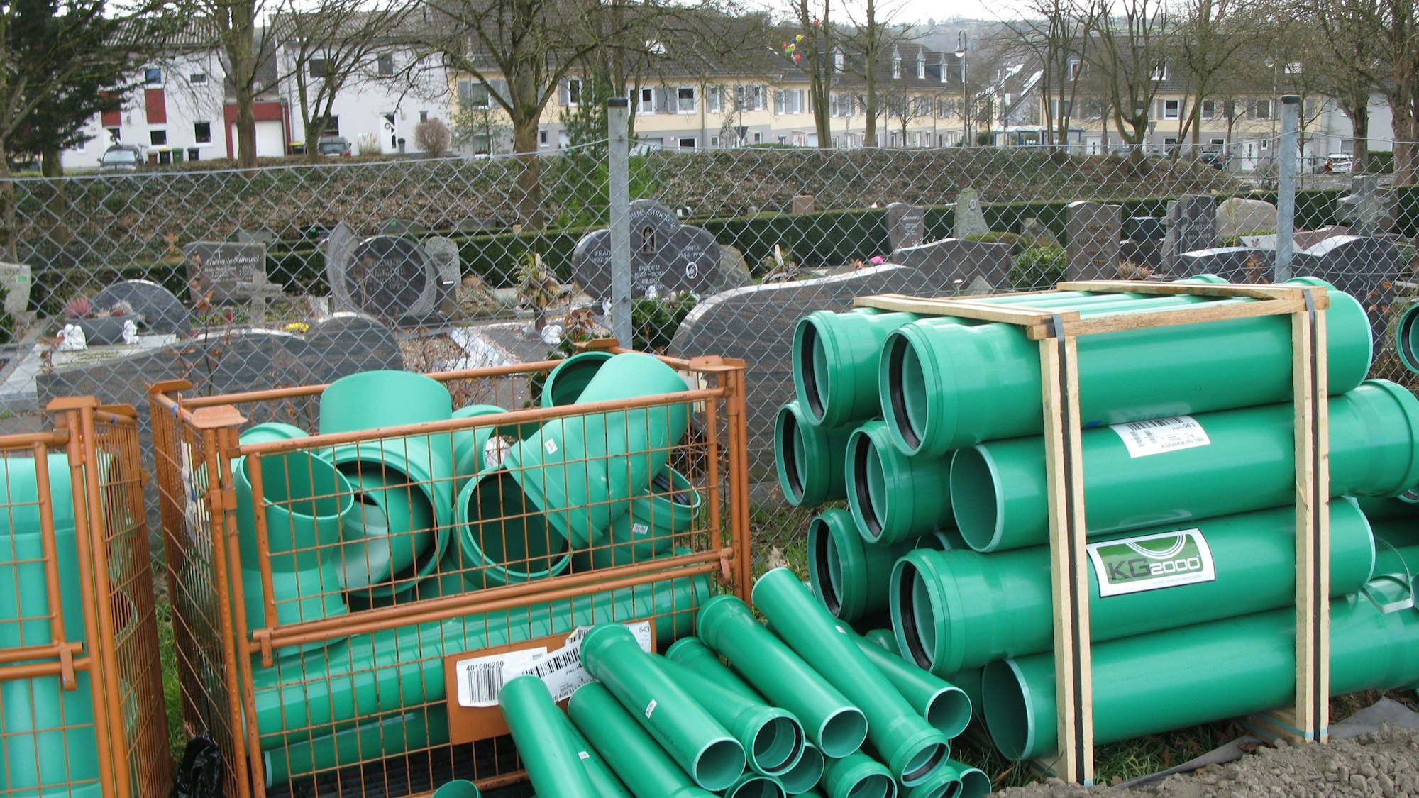 Grüne Baurohre lagern vor einem Zaun, hinter dem Grabsteine eines Friedhofs zu sehen sind.
