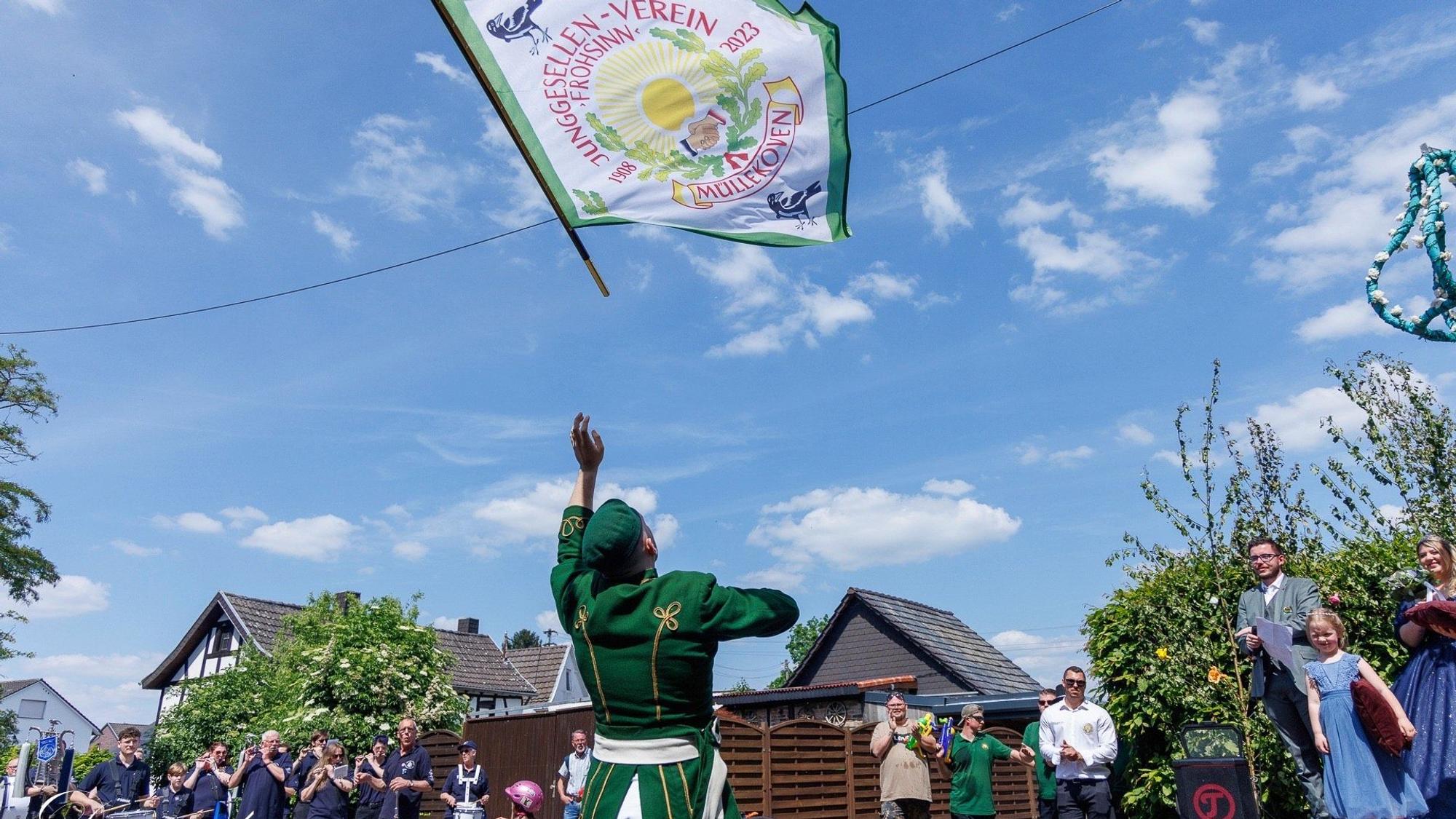 Ein Mann in militärischer Uniform hat eine Fahne in die Luft geworfen. Sie trägt die Aufschrift Junggesellenverein Müllekoven.