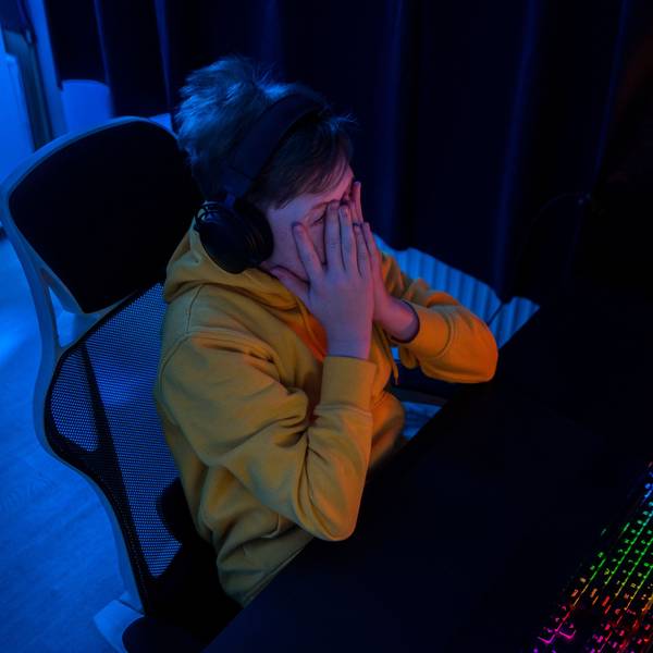 Ein Junge sitzt in einem dunklen Zimmer an einem Schreibtisch und spielt Online-Games, er hält erschrocken die Hände vors Gesicht.